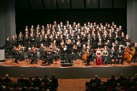 ein großer Chor steht hinter einem Orchester auf einer Konzertbühne. Im Vordergrund Dirigentin und Solisten. Ganz vorne am unteren Bildrand Publikum.